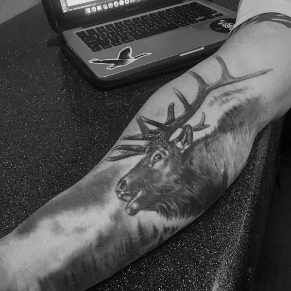 Realistic black and grey elk tattoo on the forearm, featuring detailed shading and a misty forest background for a bold and lifelike design.