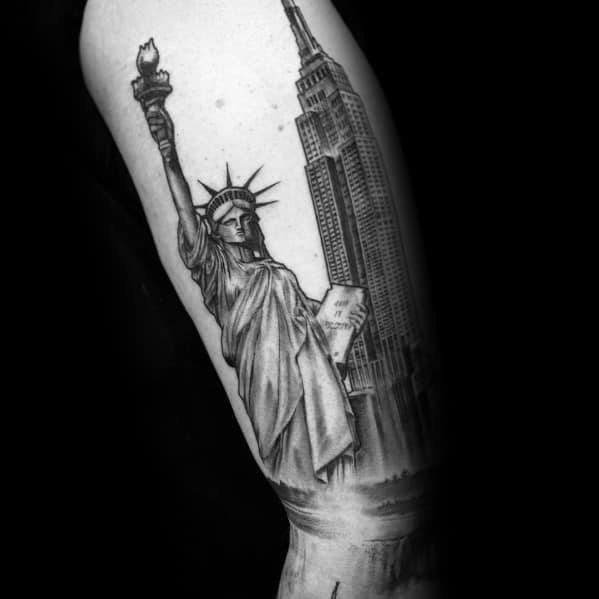 Empire State Building tattoo with the Statue of Liberty, featuring detailed shading and realism, inked on a man's upper arm.