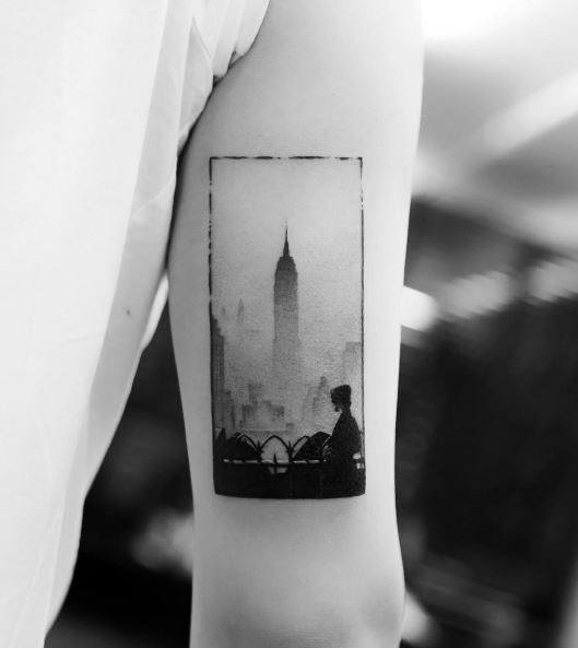 Artistic black ink tattoo of a hazy Empire State Building skyline with a silhouette figure, framed neatly on a man's upper arm.