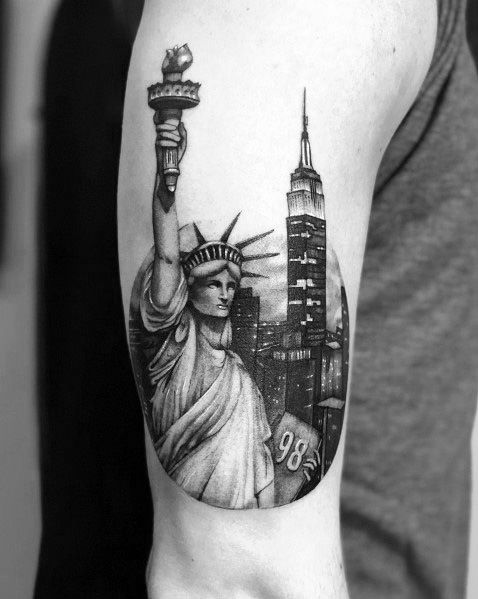 Empire State Building tattoo featuring Statue of Liberty and NYC skyline, inked with striking black shading on a man's upper arm.