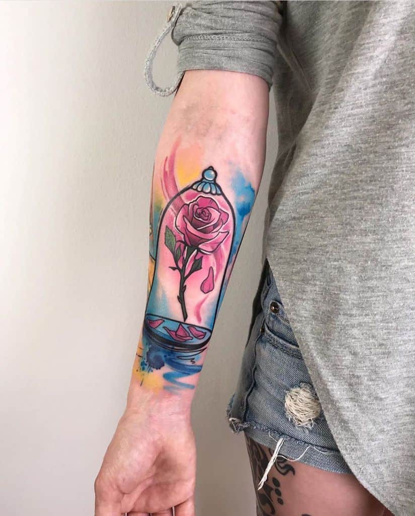 Vibrant watercolor-style forearm tattoo of the enchanted rose inside a glass dome, with bright splashes of blue, pink, and yellow hues.