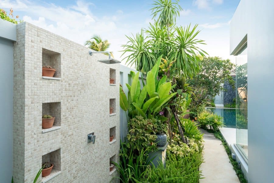 Modern outdoor shower with tiled wall, built-in planters, and lush tropical greenery near a pool.