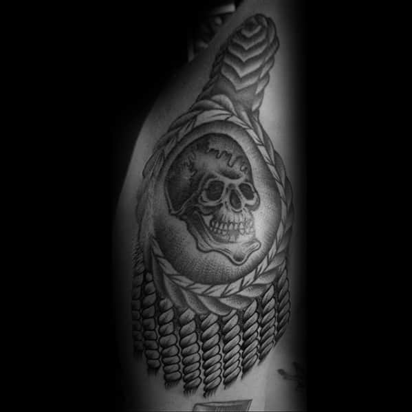 Dark-toned epaulette tattoo with a detailed skull centerpiece, thick braided border, and textured tassels flowing downward.