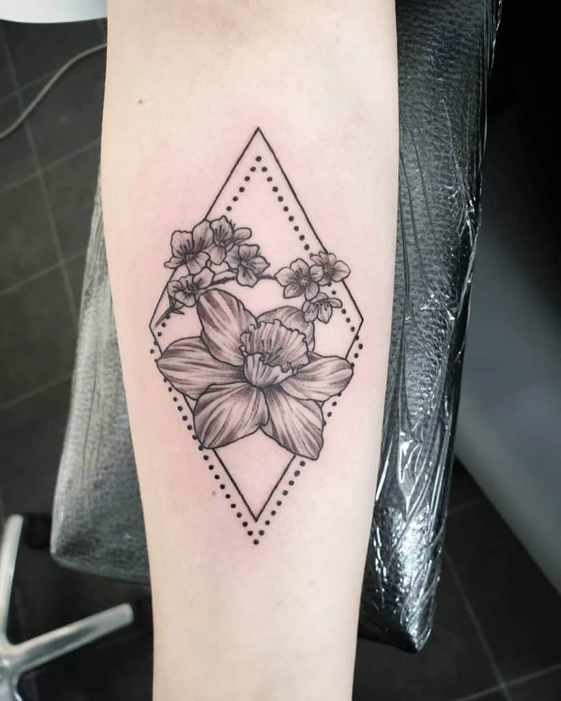 A minimalist tattoo featuring a daffodil and small flowers within a geometric diamond shape, framed by dotted lines for a clean, modern aesthetic.