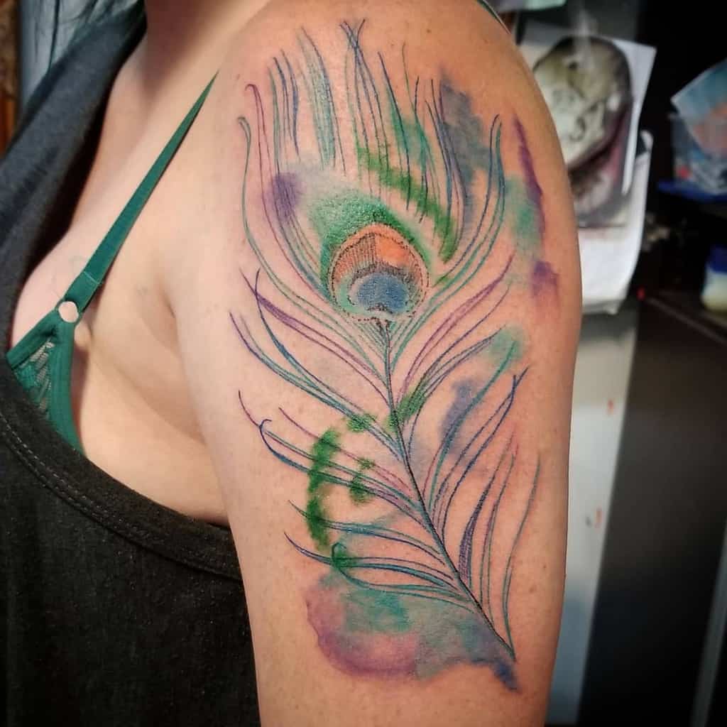 Watercolor-style peacock feather tattoo on the upper arm, featuring soft blue, green, and purple hues with delicate brushstroke effects.