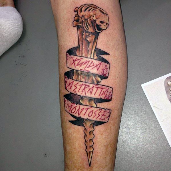 Evil Dead tattoo of the Kandarian dagger wrapped in a ribbon with Latin incantation, featuring a skull-shaped hilt and sharp detail.