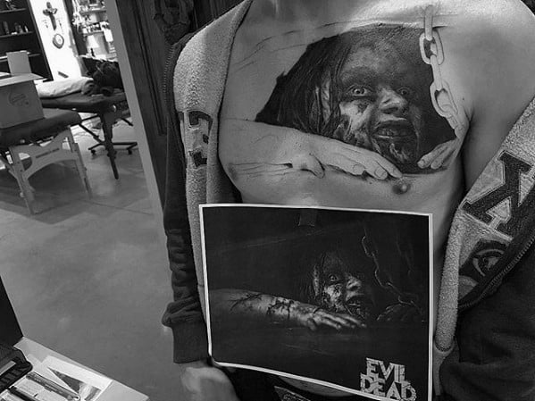 Hyper-realistic Evil Dead chest tattoo of a possessed woman in chains reaching out, replicating the iconic horror movie scene in black and grey.