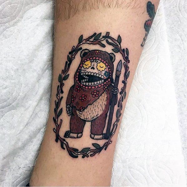 Day of the Dead-inspired Ewok tattoo with floral patterns and face paint, framed by a leafy oval border while holding a spear.