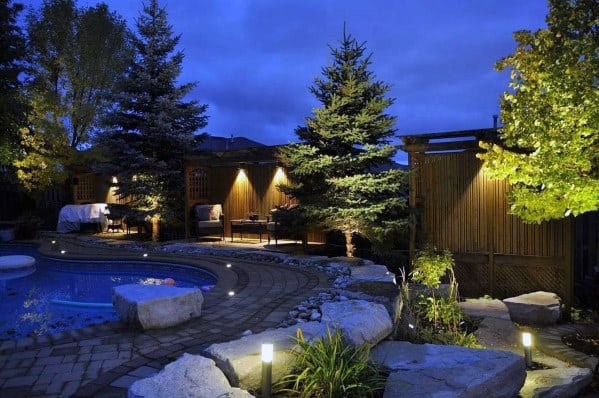 A serene backyard with a lit pool, stone pathway, and illuminated trees under a deep blue evening sky