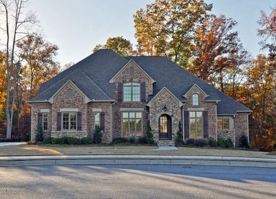 Elegant brick and stone home with symmetrical gables, arched entry, and large windows, set against a backdrop of autumn trees