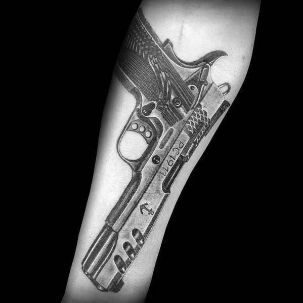 Realistic black and gray tattoo of a custom 1911 pistol on the forearm, featuring intricate details, engravings, and shading for a three-dimensional effect.