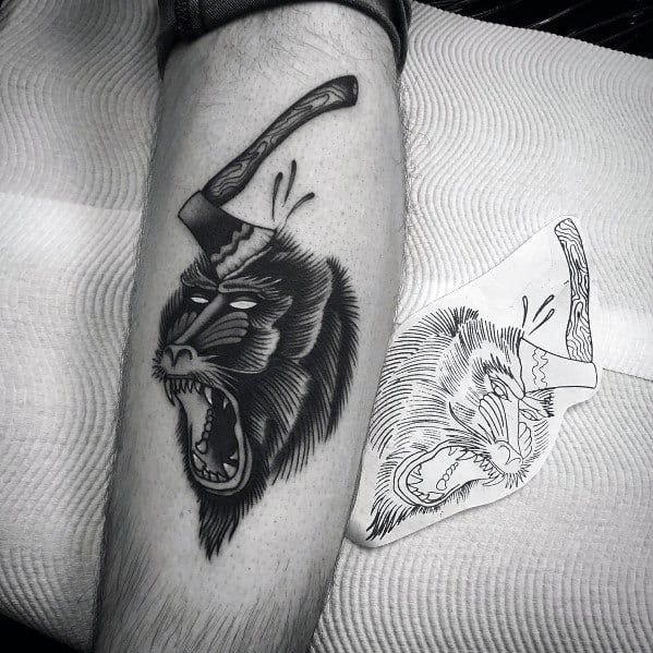 Black and grey baboon tattoo with fierce expression, bold shading, and an axe embedded in its head, creating a dramatic and intense design.