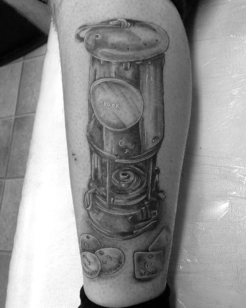 Black and grey tattoo of a detailed miner’s lamp with name tags and numbered tokens below it on the calf.
