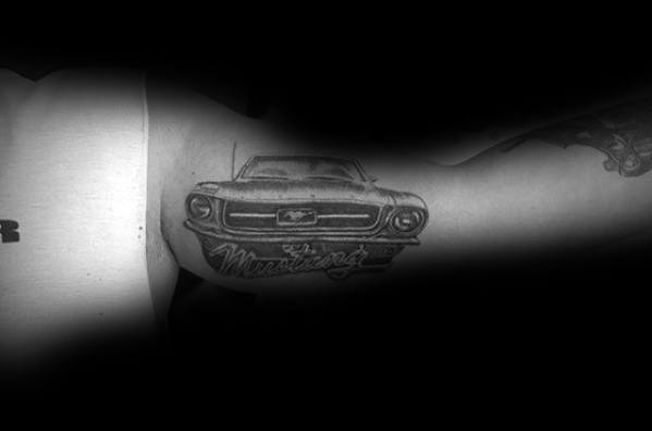 Black and grey Ford Mustang tattoo on upper arm, featuring a front view of the classic muscle car with bold lettering underneath.