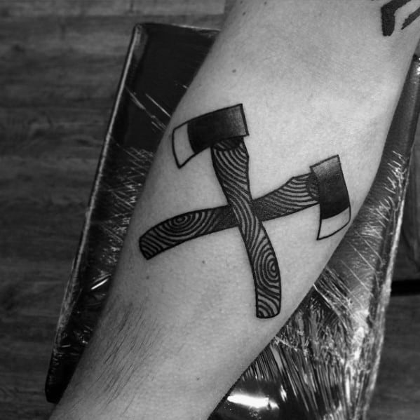 Black and grey tattoo of crossed hatchets on the forearm, featuring bold shading, wood grain texture on the handles, and sharp-edged blades.
