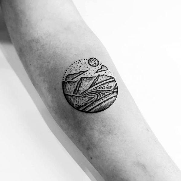 Minimalist circular mountain tattoo on the forearm, featuring a landscape with dot work details, a sun or moon, and intricate line patterns.