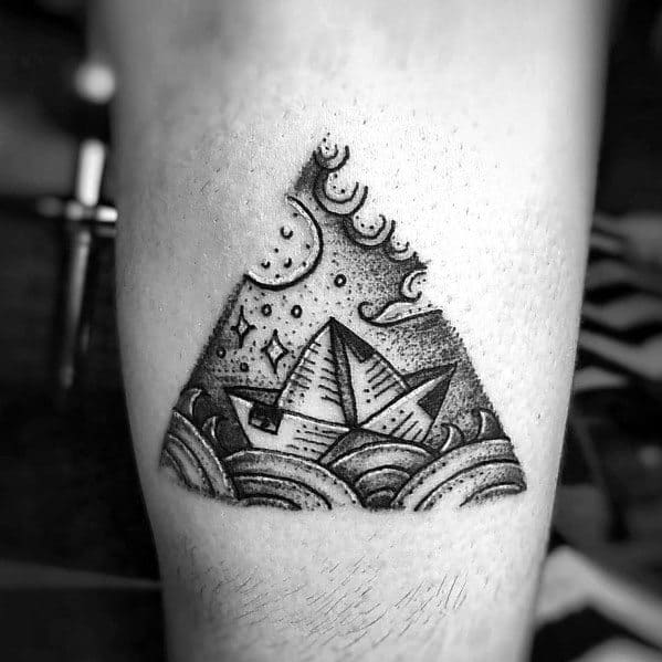Paper boat tattoo in a triangular frame with waves, moon, and stars, symbolizing adventure, dreams, and navigating life’s journey.