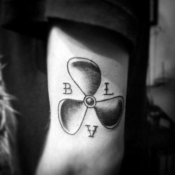 Minimalist black and grey forearm tattoo of a three-blade propeller with shaded details and the letters "B," "L," and "V" around it.