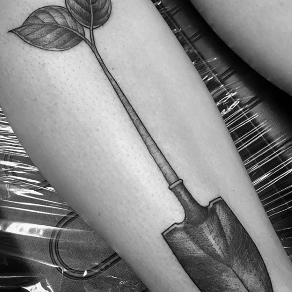 Black and grey shovel tattoo with a handle transforming into a plant stem and leaves, symbolizing growth, nature, and transformation.