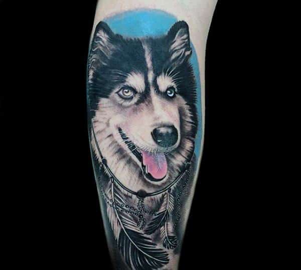 Realistic Siberian Husky tattoo on a man's leg, featuring bright blue eyes, a happy expression, and feathered accessories against a soft blue background.