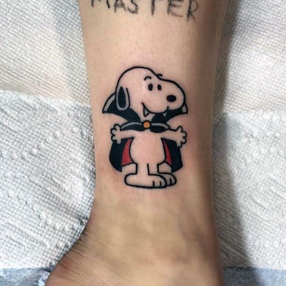 Colorful tattoo of Snoopy dressed as a vampire with a black cape, red lining, and small fangs, placed on a person's ankle.