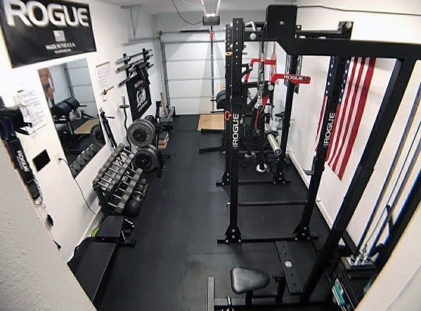 Compact home gym with Rogue equipment, weights, dumbbells, and an American flag on the wall.