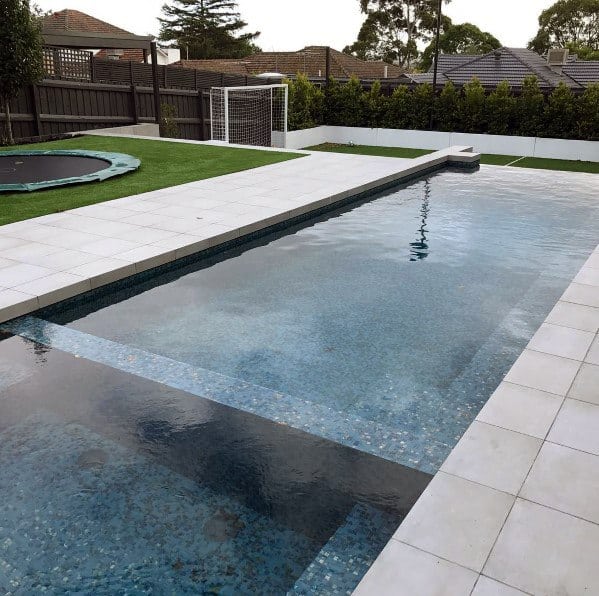 Backyard pool with mosaic tiles, white stone deck, and adjacent grass area with a trampoline.