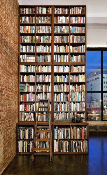 Bookshelf towers with books and a ladder, set against a brick wall and large window
