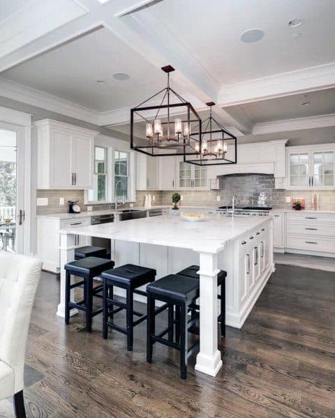 White kitchen with large island, black bar stools, dark wood floors, and modern chandeliers.