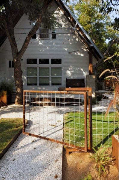 A white house with large windows, a gravel path, and an open wire fence surrounded by greenery