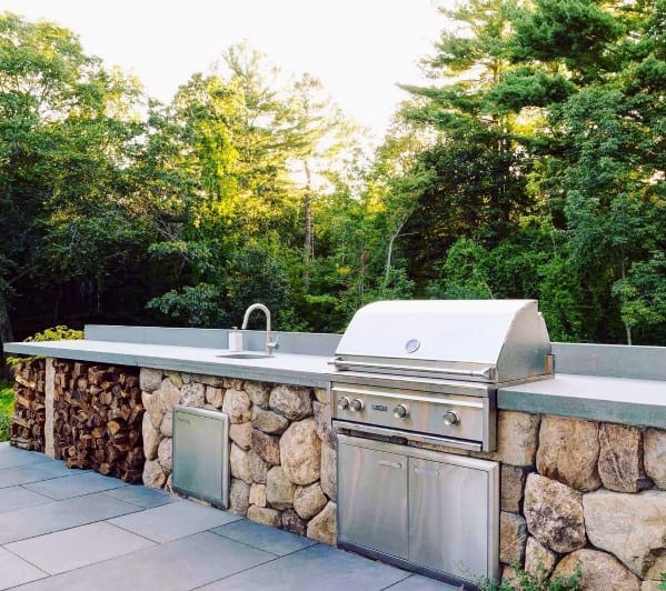 Outdoor kitchen with stone and metal, featuring a built-in grill, sink, and woodpile, set amidst lush greenery