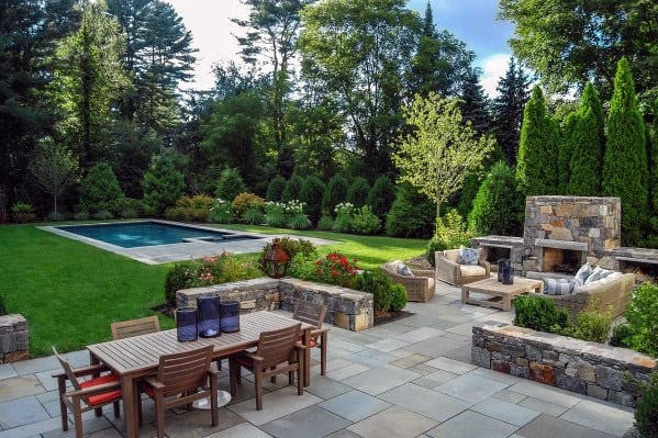 Backyard features patio seating, a dining table, stone fireplace, and swimming pool surrounded by trees and greenery