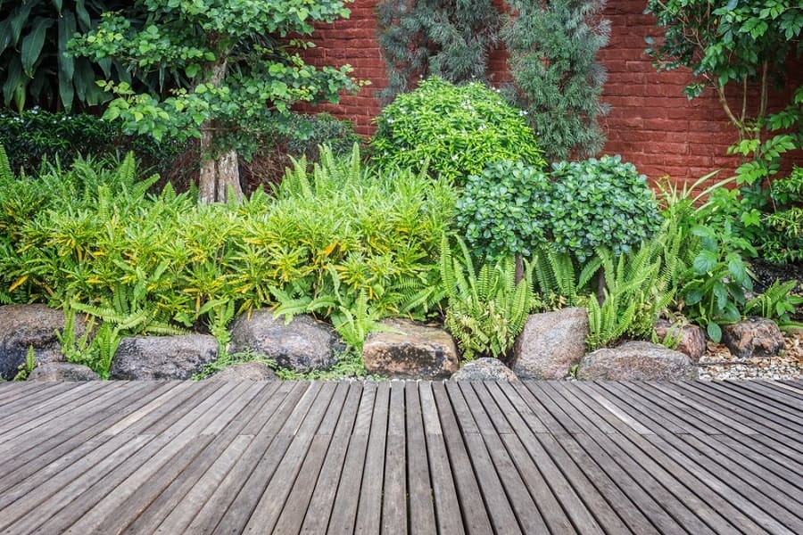 Walkway on a wooden deck before a lush garden with rocks and a red brick wall