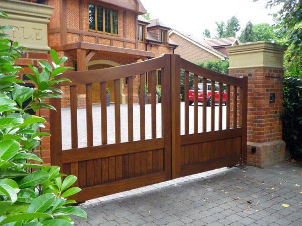Large wooden entrance gate with vertical slats, opening to a brick house and driveway with a red car parked