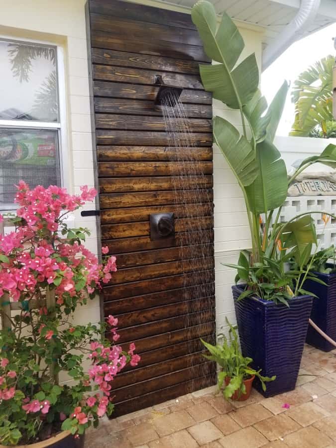 Rustic outdoor shower with dark wood slat wall, tropical plants, and vibrant pink flowers.