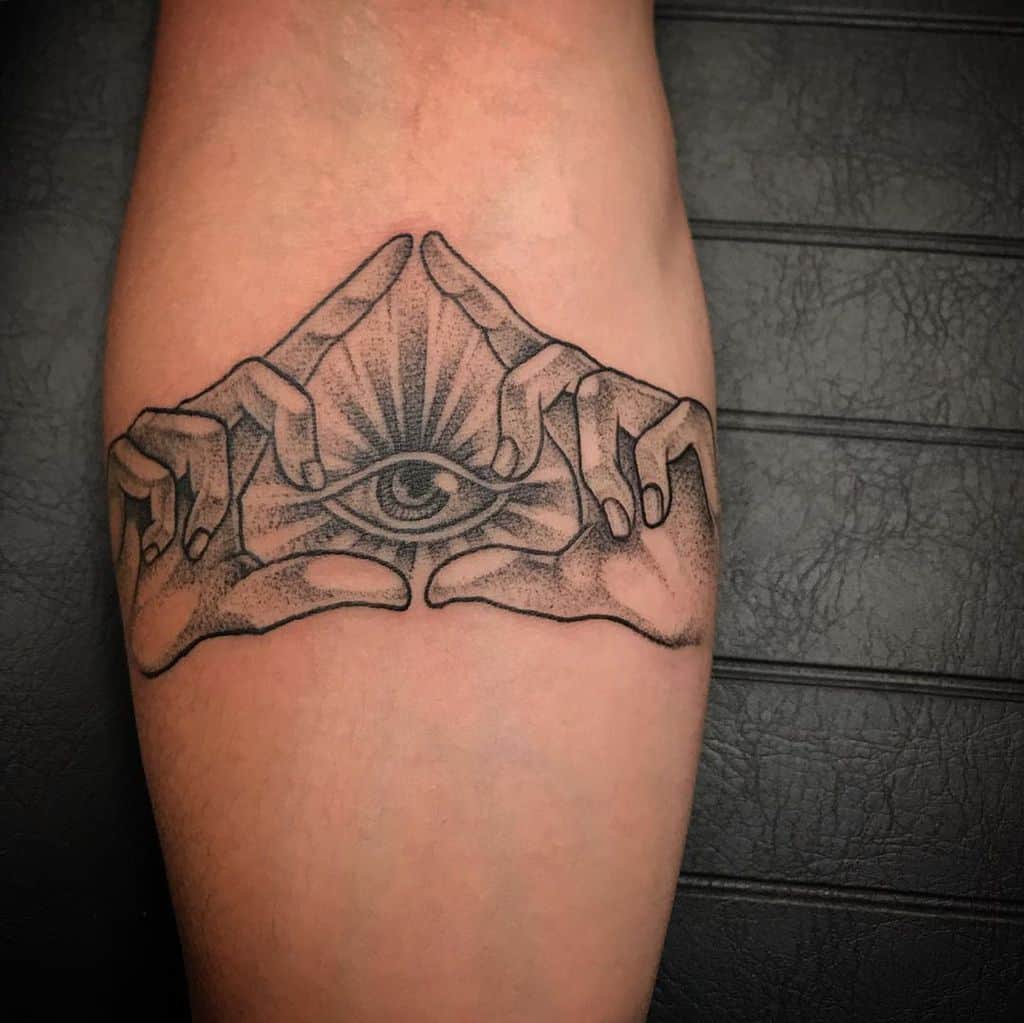 Tattoo of an all-seeing eye framed by two hands forming a triangle, with radiating lines, inked in black and dot shading on the forearm.
