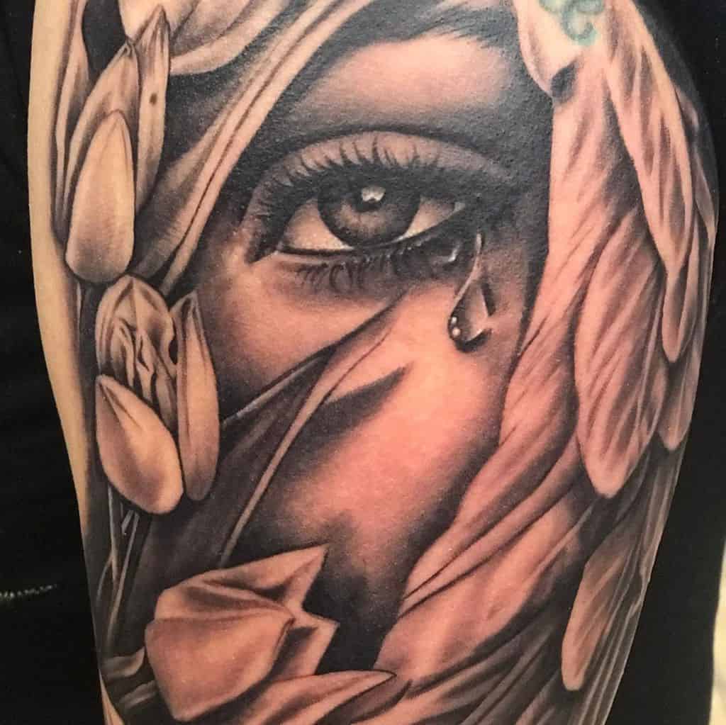 A highly detailed black and grey tattoo featuring a realistic eye with a teardrop, surrounded by tulips and flowing fabric for a dramatic effect.