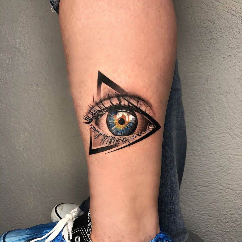 Tattoo of a highly realistic blue eye inside a black geometric triangle, inked with fine details and shading on the lower leg.