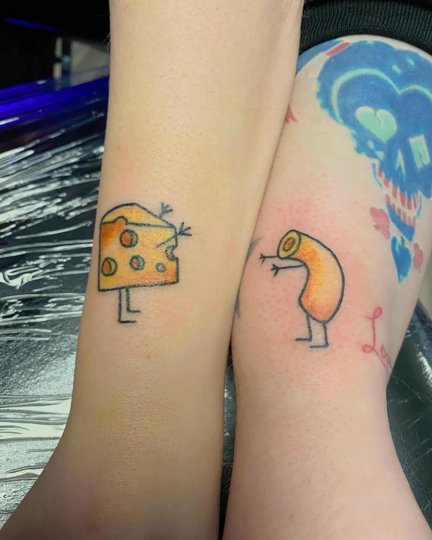 Two forearms with humorous tattoos: a cartoon cheese slice and a macaroni noodle, each with stick legs and tiny arms