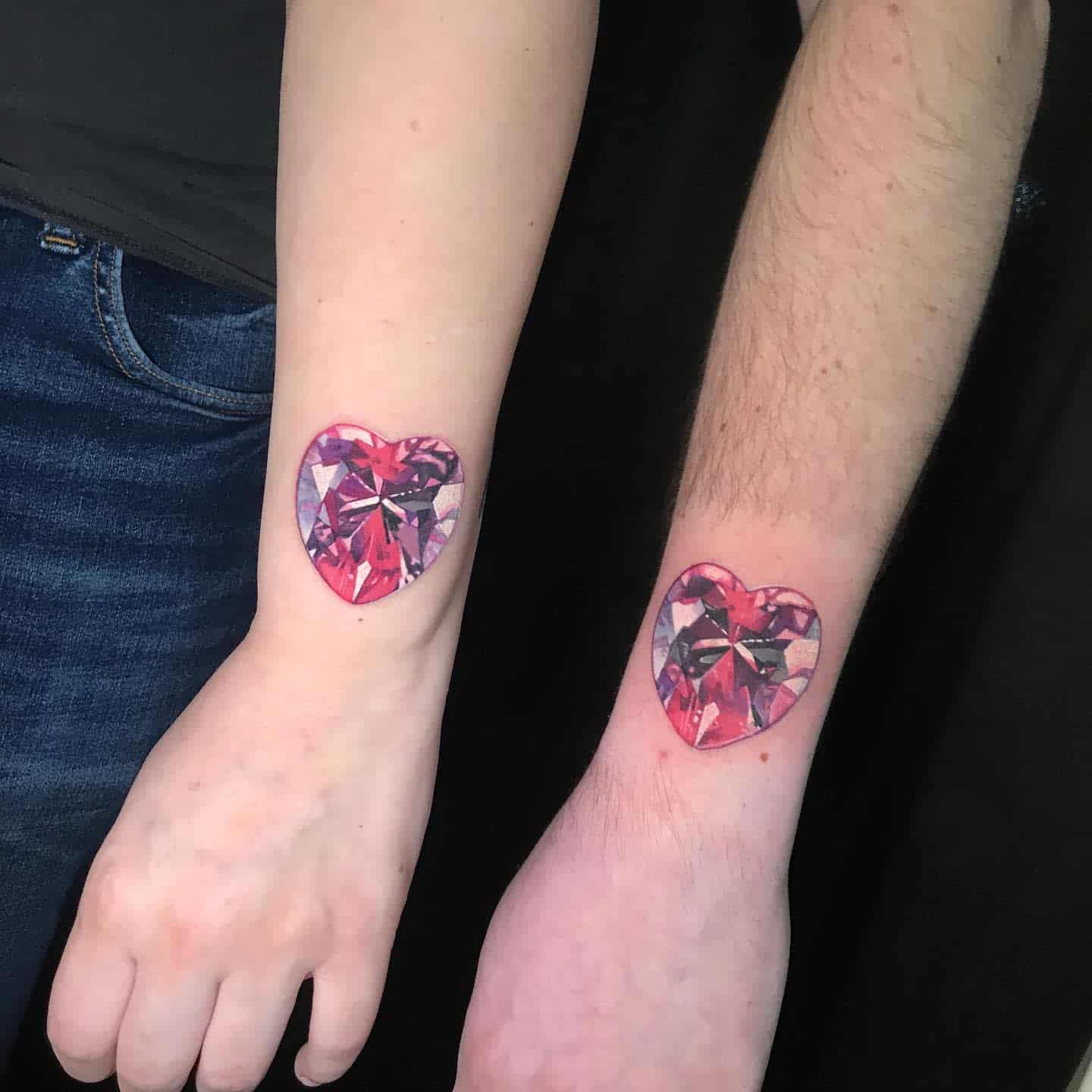Two forearms with matching heart-shaped, pink jewel diamond tattoos