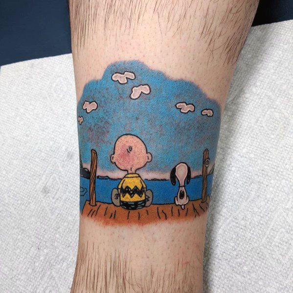Colorful tattoo of Charlie Brown and Snoopy sitting on a wooden dock, gazing at the blue sky and water with fluffy clouds.