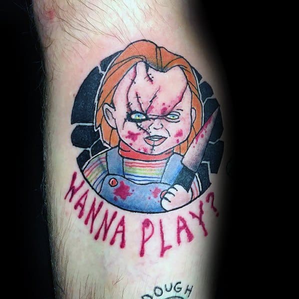 Cartoon-style Chucky tattoo with a bloodied knife, stitched face, and the phrase "Wanna Play?" in red for a bold horror-inspired design.