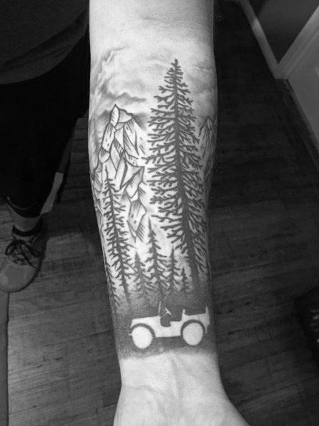 Black and grey Jeep tattoo on the forearm, featuring a classic off-road Jeep driving through a scenic mountain landscape with tall pine trees.