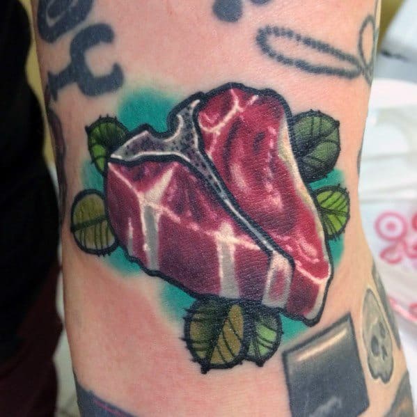 Tattoo of a detailed T-bone steak with realistic shading, surrounded by green leaves and a blue background for a bold and vibrant look