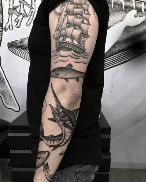 Black and grey nautical sleeve tattoo featuring a tuna fish, a ship, waves, and marine life, creating a bold and detailed ocean-themed design.