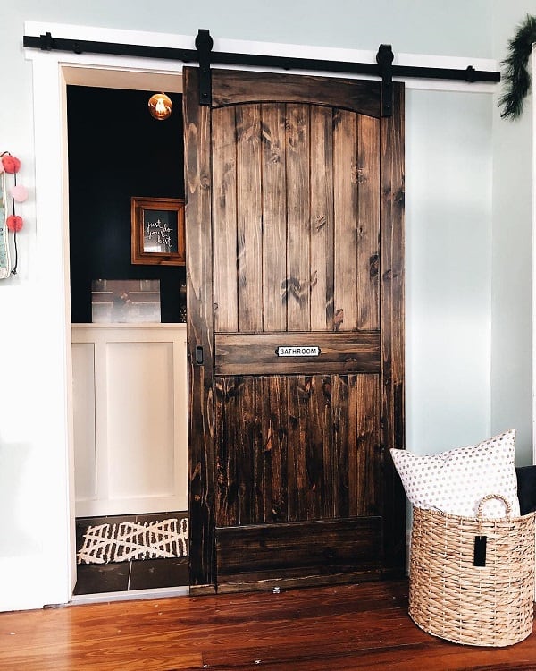 Rustic farmhouse sliding barn door with arched top leading to a bathroom with dark walls and wood accents.