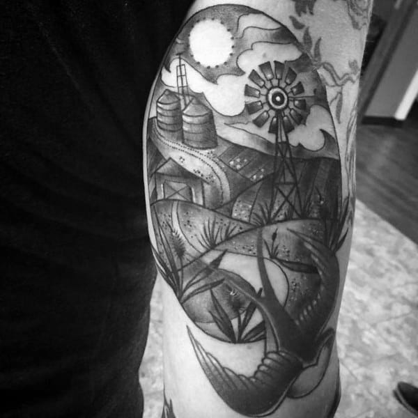 Black and gray tattoo of a rural landscape with barns, silos, windmill, and a large bird flying under a bright sun.