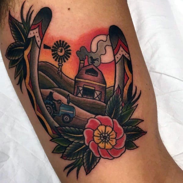Colorful traditional-style tattoo of a farm scene with a blue tractor, barn, windmill, and sunset, framed by horseshoes and flowers.