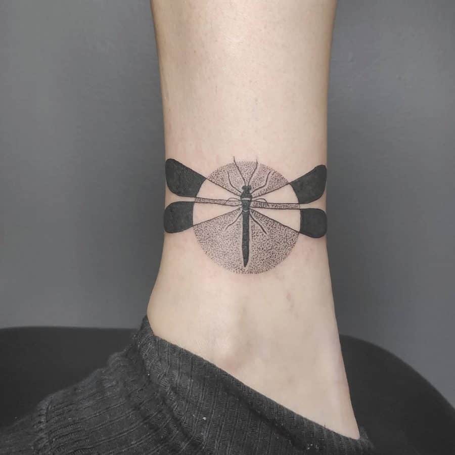 Ankle tattoo featuring a minimalist dragonfly with bold black wings and a fine dot work circle, creating a delicate yet striking design.