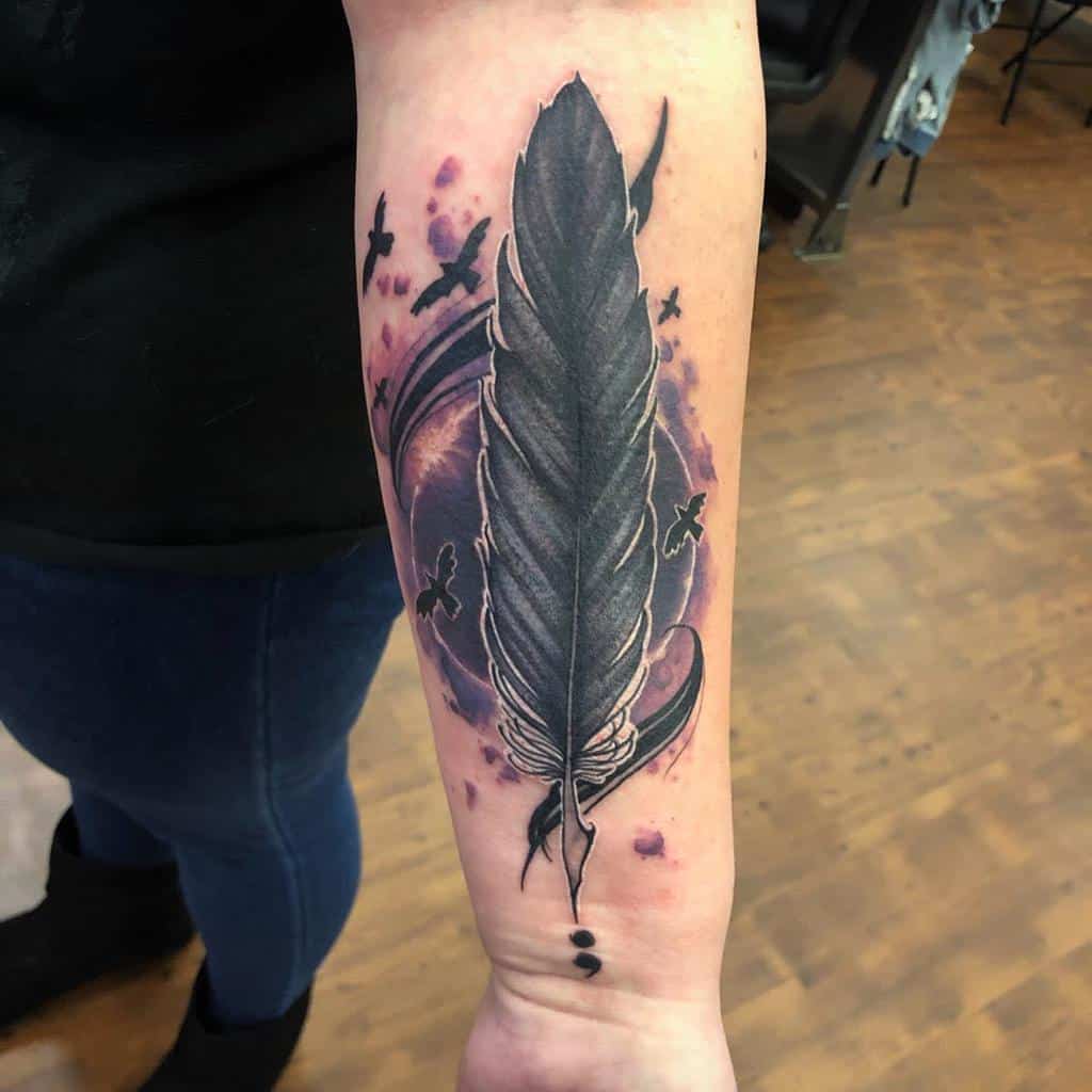 The tattoo features a detailed feather with ink splashes and birds flying around it, with a semicolon incorporated at the bottom of the feather. The design symbolizes freedom and strength.